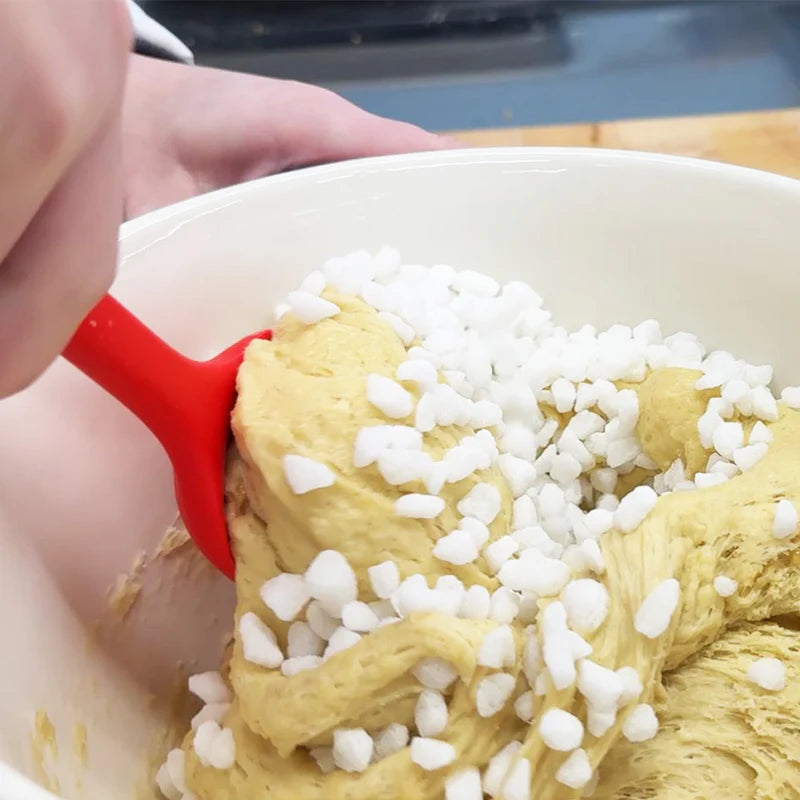 A person using a red spatula to mix dough with white pearl sugar grains in a bowl.
