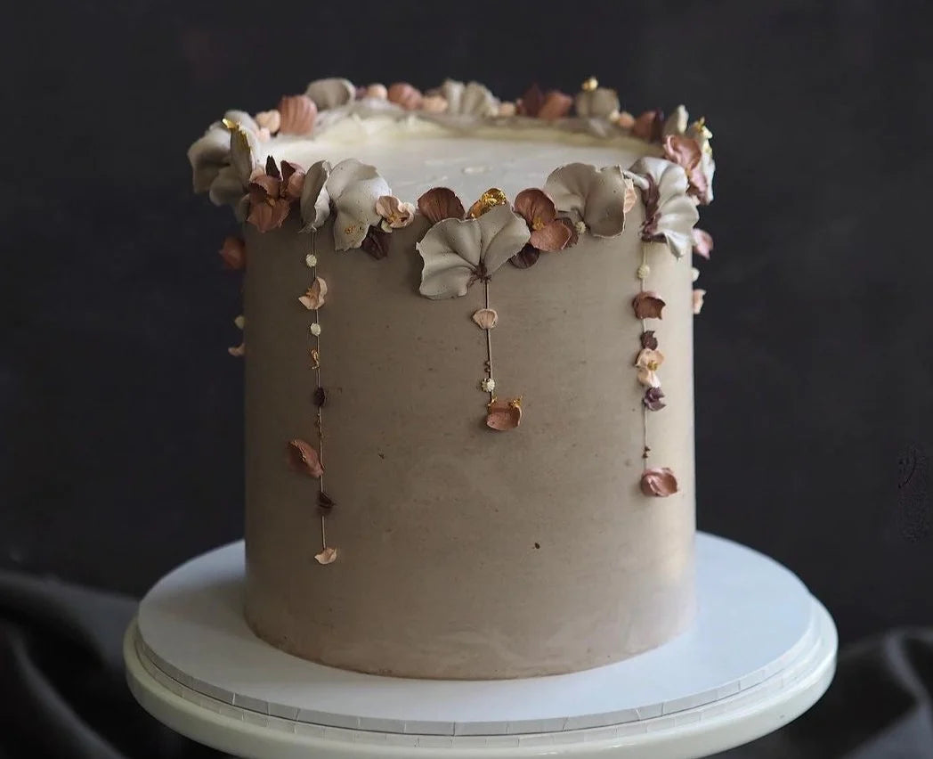 A beautifully decorated round cake sits on a white cake stand. The cake has a smooth, light brown frosting and is adorned with delicate, softly colored edible flowers in shades of beige, pink, and gold. Some flowers have small golden accents, and there are also tiny beads and floral embellishments cascading down the sides of the cake. The background is a dark, muted tone, which highlights the cake&