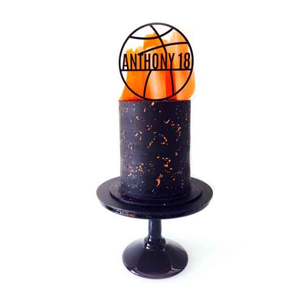 A round black cake with orange sprinkles is displayed on a black cake stand. The cake is topped with orange edible decorations that resemble flames. Above the cake, there is an acrylic cake topper in the shape of a basketball, featuring the name &quot;ANTHONY&quot; and the number &quot;18&quot; inside.