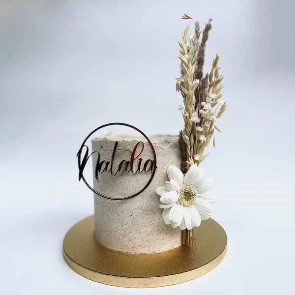 A round cake with a textured, light beige frosting is displayed on a gold circular cake board. The cake features an elegant acrylic decoration with the name &quot;Natalia&quot; inside a circular frame. Adorning one side of the cake are dried flowers arranged alongside a white daisy, adding a natural touch to the design. The background is a soft, neutral color, highlighting the cake&