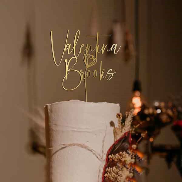 A close-up of a wedding cake with a personalized cake topper. The topper features the name &quot;Valentina Brooks&quot; in an elegant, cursive gold font, accompanied by a simple flower illustration. The cake appears white with a textured finish, and there are decorative elements, including dried flowers, surrounding the base. The background is softly blurred with warm tones and hanging lights, creating a romantic atmosphere.