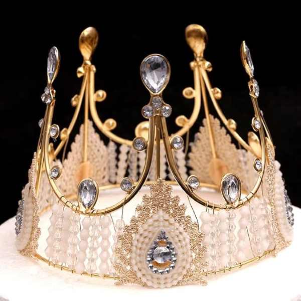 A decorative cake topper designed to resemble a tiara or crown. It features a golden metallic frame with intricate detailing, including several peaks adorned with clear gemstones and pearl-like elements. The base has a lace-like pattern, embellished with sparkling crystals and beads. The overall look is elegant and royal, suitable for celebratory cakes or special occasions.