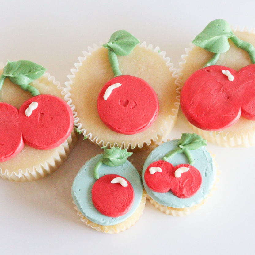 A collection of decorative cupcakes arranged on a white surface. The cupcakes are topped with colorful frosting designed to resemble cherries. Two cupcakes feature a pair of red cherry shapes, with green leaves, while another has a blue background with a similar cherry design. The overall appearance is bright and appealing, highlighting the use of food coloring in the cupcake decorations.