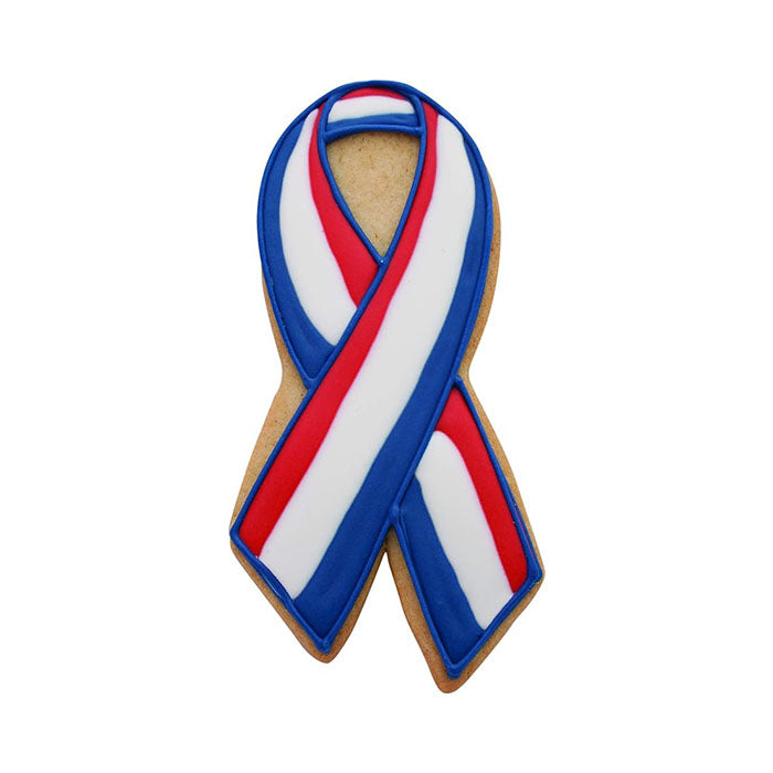 The image shows a cookie shaped like an awareness ribbon. The cookie is decorated with red, white, and blue icing in a striped pattern. The ribbon design features a blue border, a wide white stripe in the center, and thinner red stripes on both sides. The cookie has a golden-brown color base.