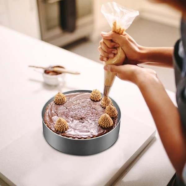 A person is decorating a chocolate cake with a piping bag. The cake, which has chocolate frosting, is placed on a white countertop. The person is adding decorative swirls or peaks on top of the cake using a light brown frosting, with several similar decorations already in place. In the background, there?s a small bowl with a spoon, likely containing more frosting or ingredients. The setting appears to be a kitchen.