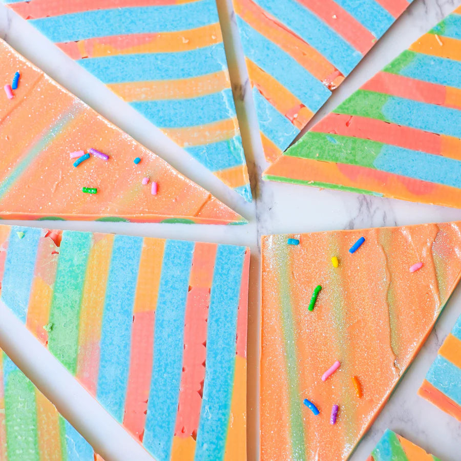 The image shows a beautifully arranged plate of colorful, triangular slices of a dessert. Each slice features a vibrant pattern of alternating stripes in shades of sky blue, pink, orange, and green. Some slices are adorned with small, colorful sprinkles scattered on top, adding to the festive and playful appearance. The background is a light-colored surface, highlighting the bright colors of the dessert slices.