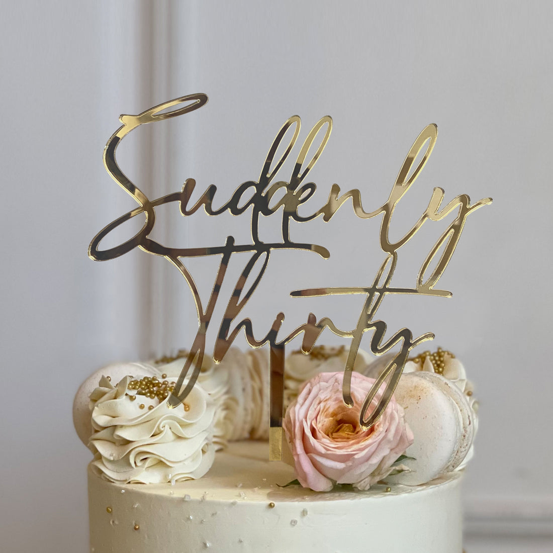 An elegant cake with a smooth, light-colored frosting is topped with a customized acrylic cake topper that reads "Suddenly Thirty" in a stylish, cursive font. The topper is golden and stands prominently above the cake. Surrounding the topper, the cake features decorative elements such as swirls of cream, edible gold pearls, a pink rose, and white macarons. The overall presentation suggests a celebratory theme, suitable for a 30th birthday party.