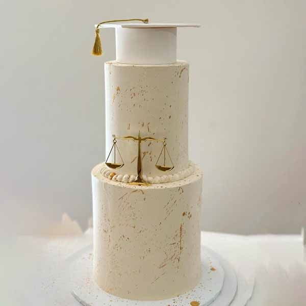 A three-tiered white cake with a textured finish, featuring a small graduation cap on the top tier, complete with a golden tassel. The middle tier is adorned with a decorative rope border and displays a gold scale of justice, symbolizing balance. The cake sits on a round white cake board, and subtle gold accents are scattered across the surface of the cake for an elegant touch.