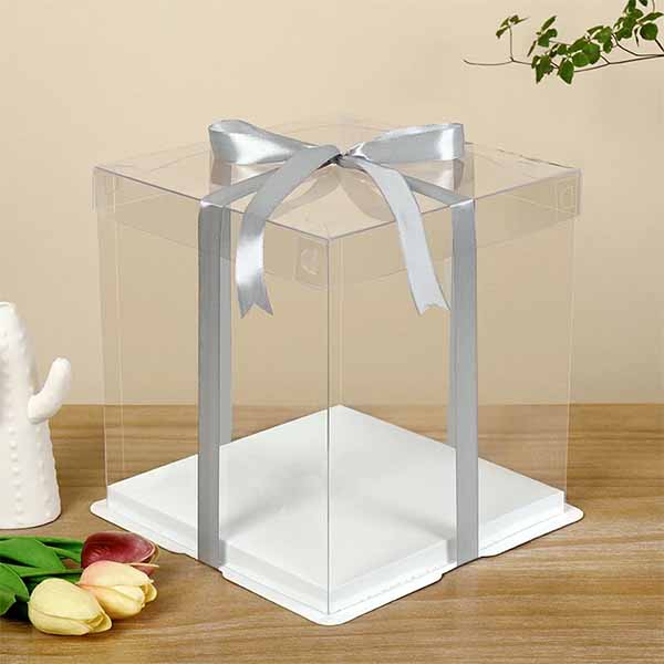 A square clear lid cake box, measuring 8.5 inches wide and 11.8 inches tall, is displayed on a wooden surface. It features a transparent design with a silver ribbon tied in a bow around it. The base of the box is white, and there are colorful tulips and a decorative ceramic piece nearby. The background consists of a simple, neutral-toned wall.