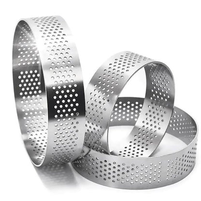 Three stainless steel perforated tart rings are displayed, each with a shiny finish. The rings are circular, featuring evenly spaced holes throughout their surfaces. They vary slightly in size, with the largest ring having a diameter of approximately 10 cm and a height of about 2.5 cm. The rings are positioned in a way that showcases their round shapes and perforated designs.