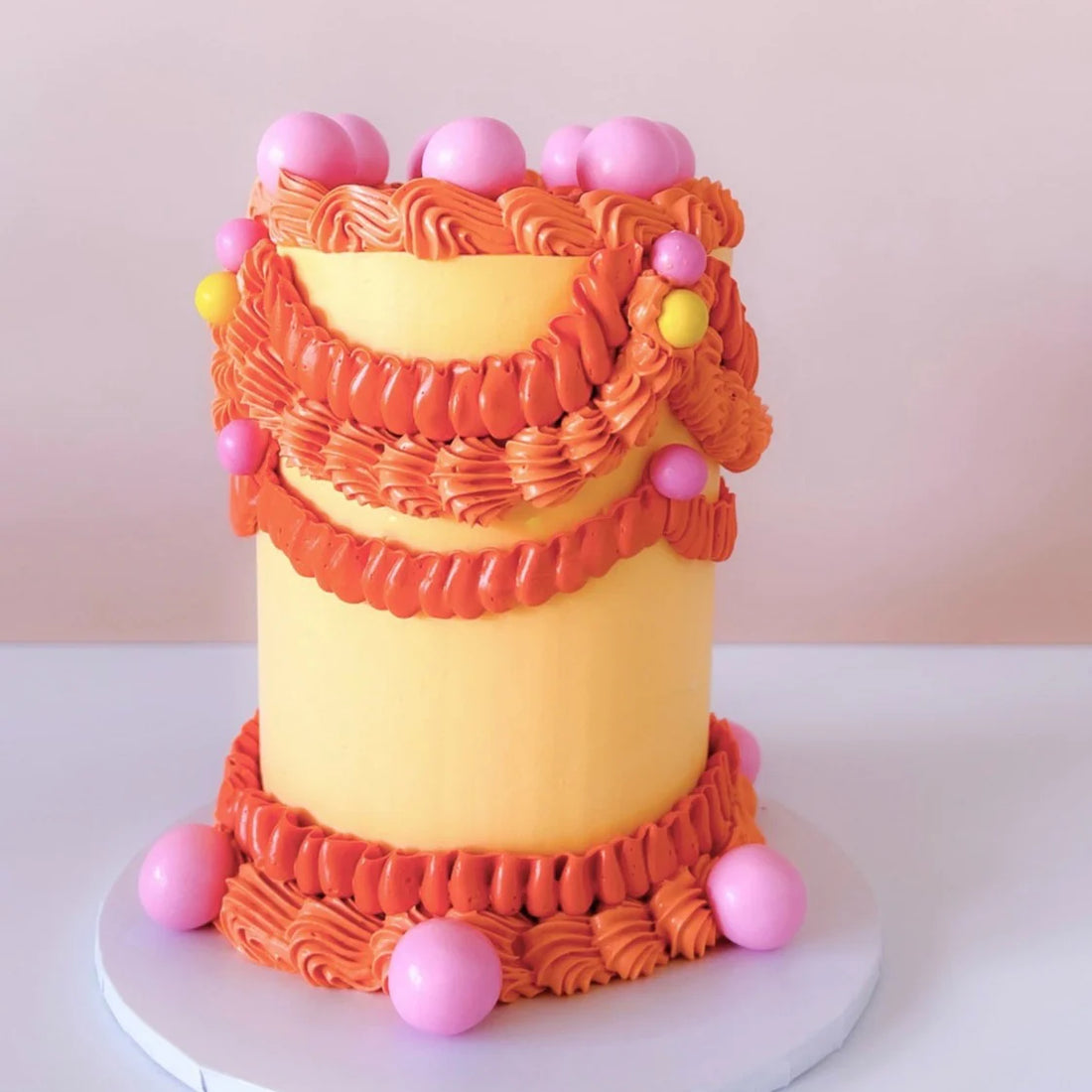 A vibrant, decorated cake with a yellow frosting base featuring multiple layers of orange and pink icing swirls. The cake is adorned with colorful round decorations, including orange, pink, and yellow balls positioned around its sides and on top. It is set on a light grey round cake board against a soft pink background.