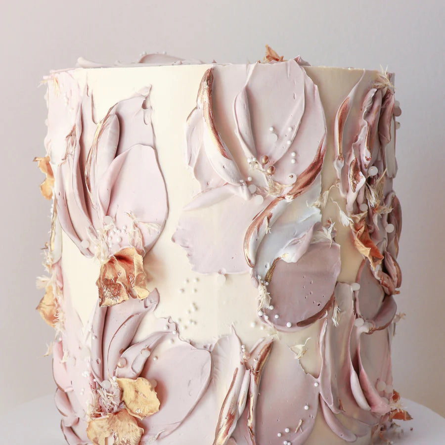 A decorated cake is shown, featuring a textured design with pastel colors. The cake is primarily white with intricate floral patterns made from icing in tones of taupe and soft pink. Some flowers have golden accents, and small decorative elements like white pearls are scattered throughout the design. The background is neutral, allowing the cake?s details to stand out.