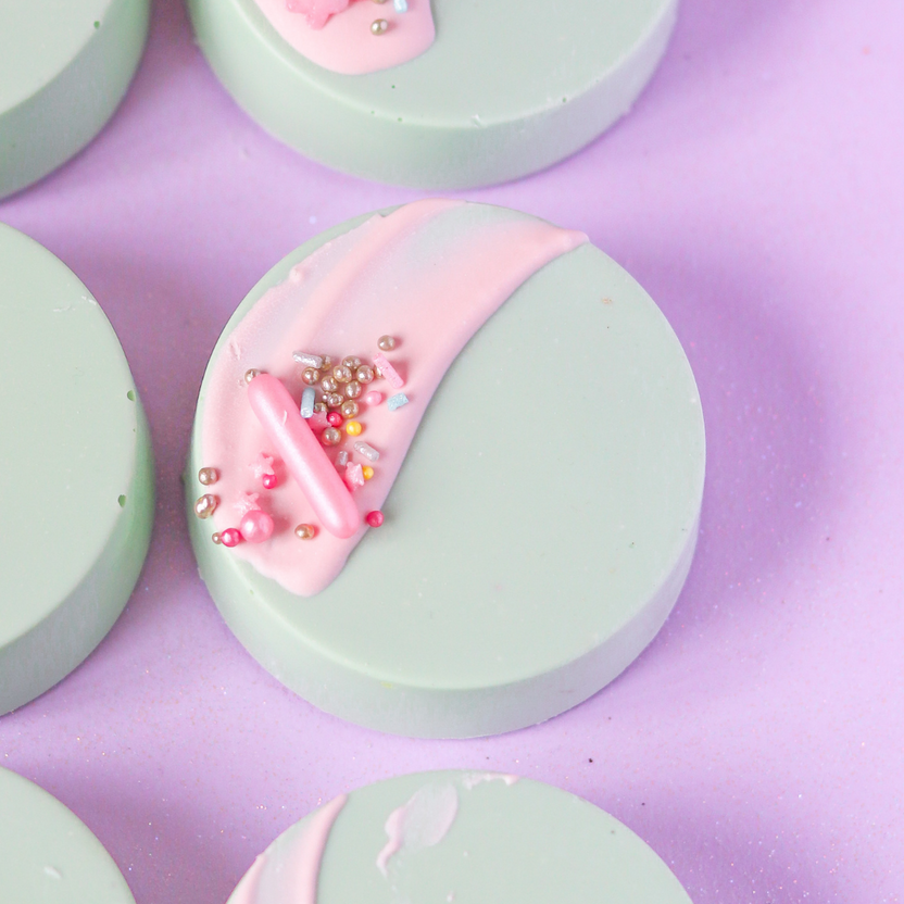 A close-up image of round, mint green discs with decorative elements. One disc features a streak of pale pink icing and various colorful sprinkles on top. The background is a soft pink surface, enhancing the pastel colors of the discs and decorations.