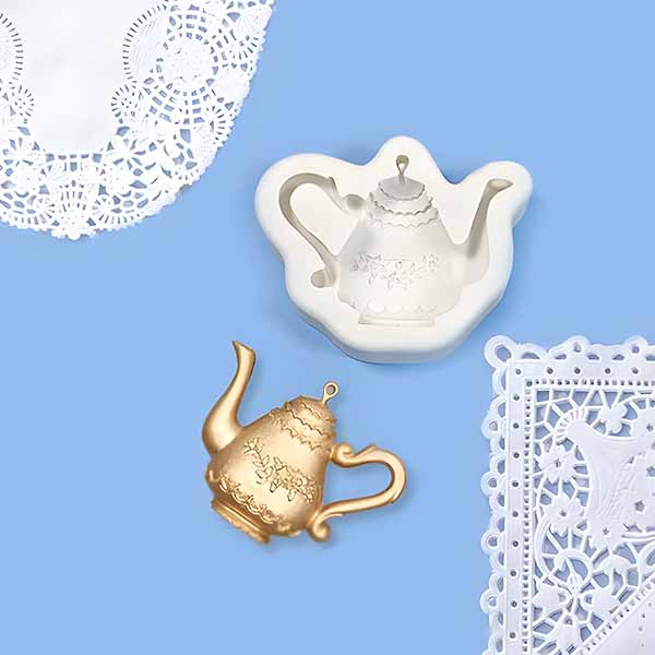 A flat lay showing a white silicone mould shaped like a teapot, alongside a small gold teapot-shaped object. The background is light blue, and there are decorative lace doilies positioned partially in the frame, one white doily on the left and another intricate white piece on the right.