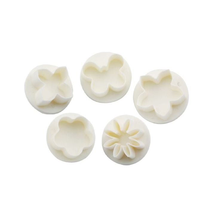 Five white plastic plunger cutters arranged in a circular shape, each designed to create flower shapes. The cutters vary in design, featuring different floral outlines including a simple blossom, a five-petaled flower, and another with more intricate petal details. They are placed on a flat surface, showcasing their clean edges and uniform size, ideal for crafting or baking purposes.