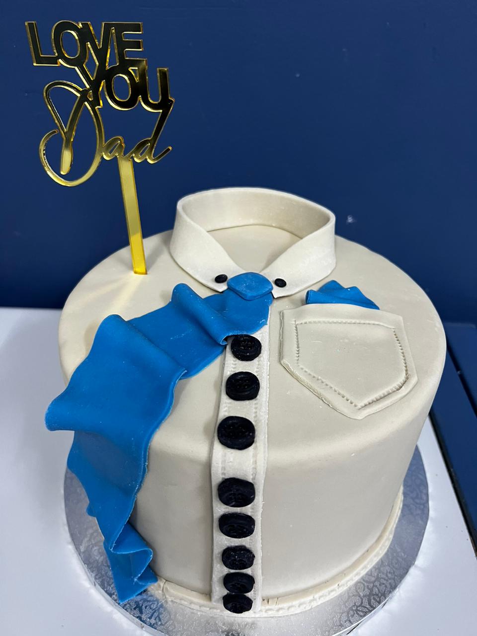 A round cake designed to resemble a dress shirt is displayed. The cake is covered in a smooth, light-colored frosting. It features a white collar and a blue fondant ruffle that runs diagonally across the front. There are black fondant buttons arranged in a line down the center, with a small pocket detail on one side. Topping the cake is a gold acrylic cake topper that reads &quot;LOVE YOU DAD.&quot; The cake is placed on a silver, decorative cake board against a blue background.