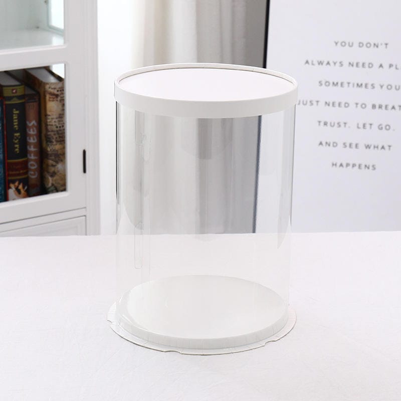 A clear, round PVC cake box with a white top and base. The box is designed to store cakes and has a cylindrical shape, measuring approximately 11.8 inches in diameter and 17.7 inches in height. It is placed on a flat, light-colored surface, with a softly blurred background showing part of a bookshelf and wall decor featuring text.