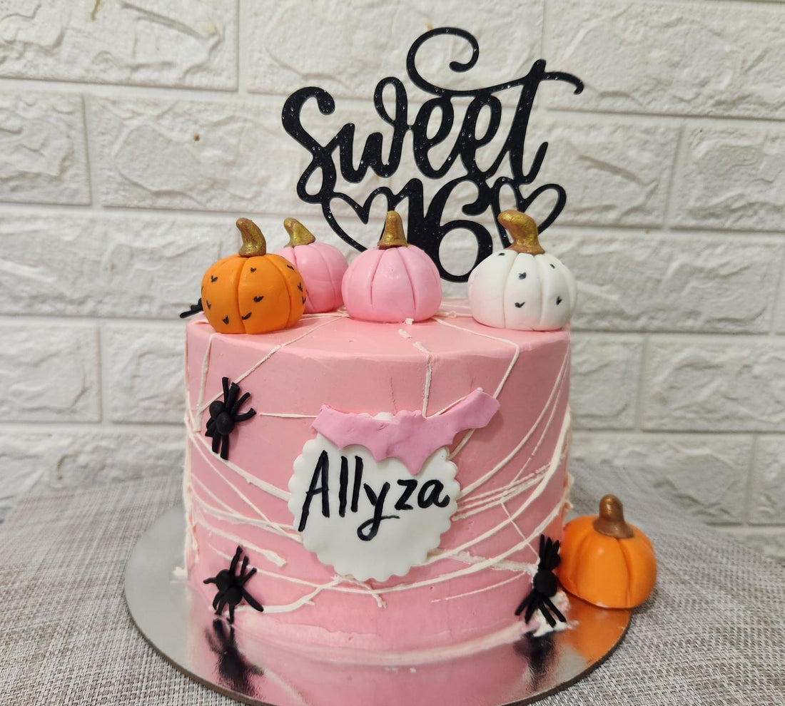 A tiered pink birthday cake adorned with a black cardstock topper that says "Sweet 16." The cake is decorated with small fondant pumpkins in orange and white, and has black spider decorations on the sides. A pink flag with the name "Allyza" is attached to the front of the cake. The background features a light-colored, textured wall.