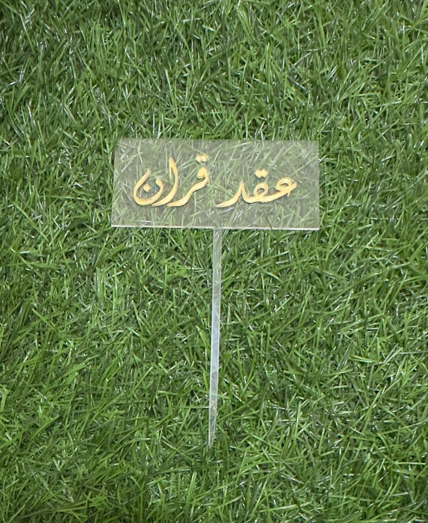 A clear acrylic cake topper with a rectangular shape, featuring elegant gold lettering that reads in Arabic. It is mounted on a thin stick for easy placement on a cake. The topper is set against a backdrop of artificial green grass.