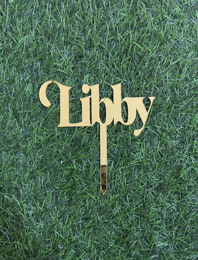 A gold acrylic cake topper featuring the name &quot;Libby&quot; in an elegant cursive font. The topper is placed on a background of green grass, highlighting its shiny surface and design. The topper has a pointed base for easy insertion into a cake.