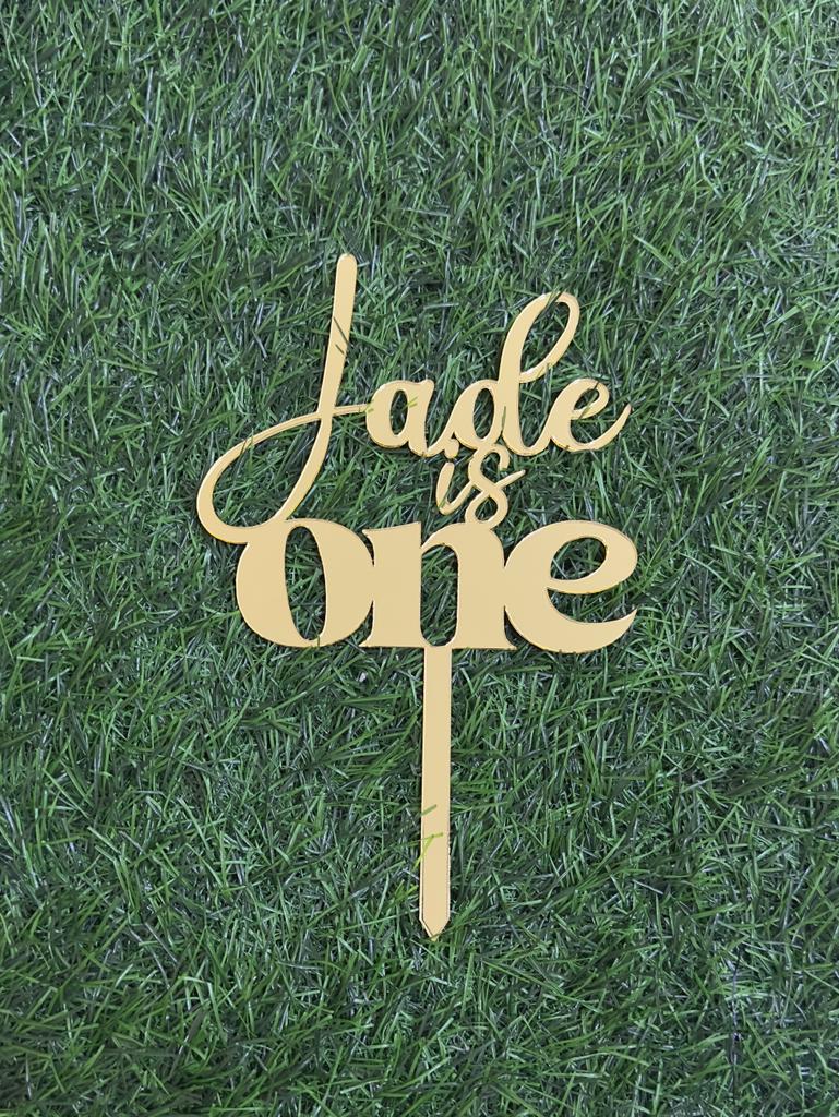 A gold acrylic cake topper is displayed on a green grass-like surface. The topper features the text &quot;Jade is One&quot; in an elegant, flowing script, with &quot;Jade&quot; in a larger font size than the rest of the text. The design includes a stake for inserting into a cake.