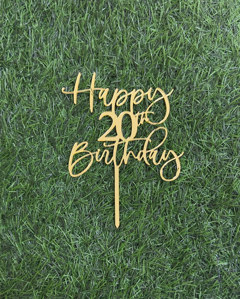 A golden acrylic cake topper featuring the phrase &quot;Happy 20th Birthday&quot; in a decorative, cursive font. The topper is placed on a background that resembles green grass, highlighting its design.