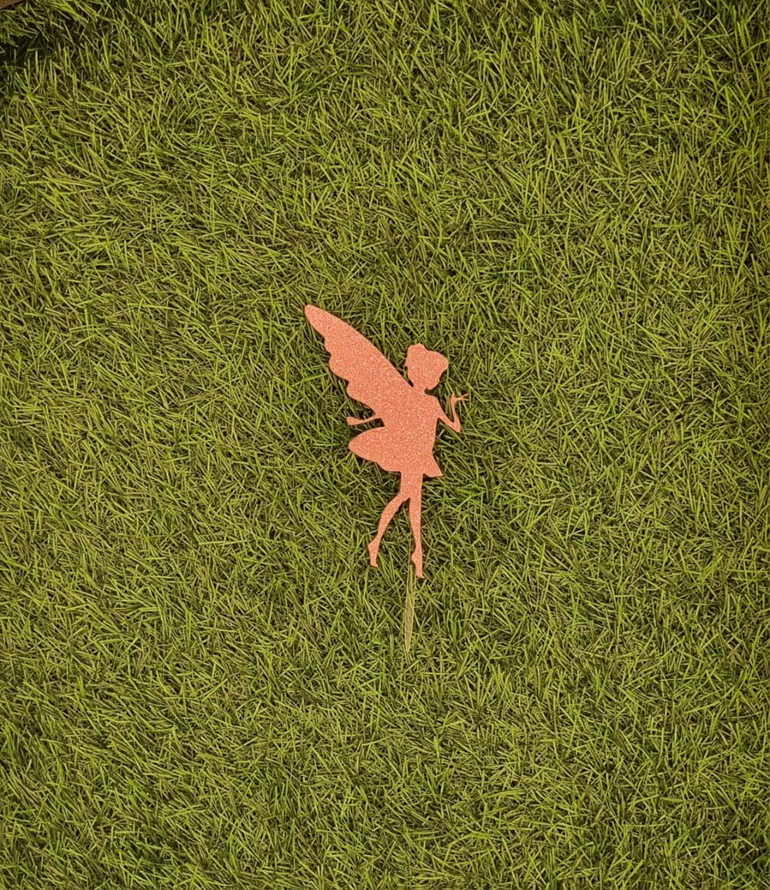 A fairy cake topper is displayed on a grassy background. The topper features a silhouette of a fairy with outstretched wings, crafted from a shimmering orange material. The fairy is posed with one hand raised, and the design includes delicate details like a flowing dress and pointed shoes. The topper is mounted on a pointed base for easy insertion into a cake.