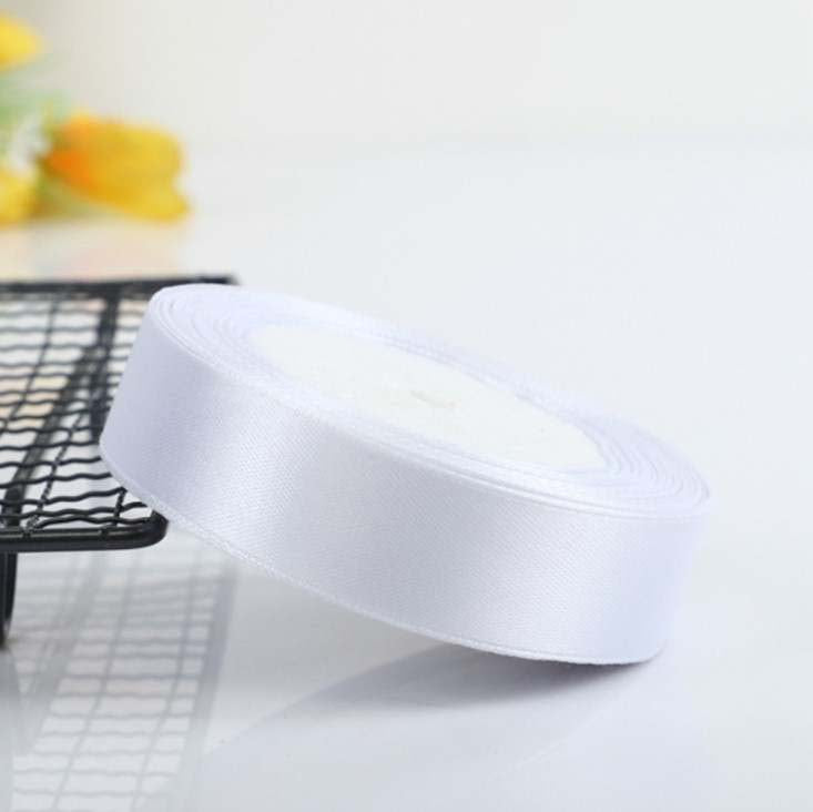 A roll of white ribbon is displayed on a black wire rack. The ribbon has a shiny, satin finish and is approximately 2.5 cm in width. The background is light and neutral, with a hint of yellow flowers visible in the blurred background, adding a soft touch to the overall image.