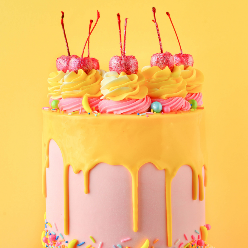 A colorful cake with a pink base is topped with yellow drippy icing and decorative swirls in yellow and pink frosting. The cake is adorned with three sparkling red cherries on top and is sprinkled with colorful decorations, including small candies and sprinkles. The background is a bright yellow, enhancing the cheerful appearance of the cake.