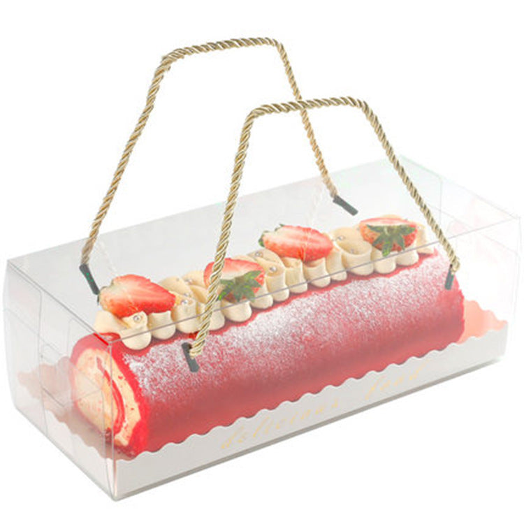 The image features a clear, rectangular dessert box with two decorative handles. Inside the box is a visually appealing Swiss roll cake, characterized by a vibrant red outer layer and a creamy filling. The top of the cake is adorned with small meringues and fresh strawberries, adding a touch of elegance. The box is slightly open at one end, allowing a view of the Swiss roll, which is dusted with powdered sugar. The overall presentation highlights the dessert as a delightful and attractive treat.