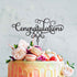 A decorated cake featuring a black acrylic topper that reads "Congratulations" in elegant cursive. The cake is adorned with macarons, strawberries, and various fresh flowers, creating a festive and celebratory appearance. The background is light and neutral, emphasizing the colorful cake and decorations.
