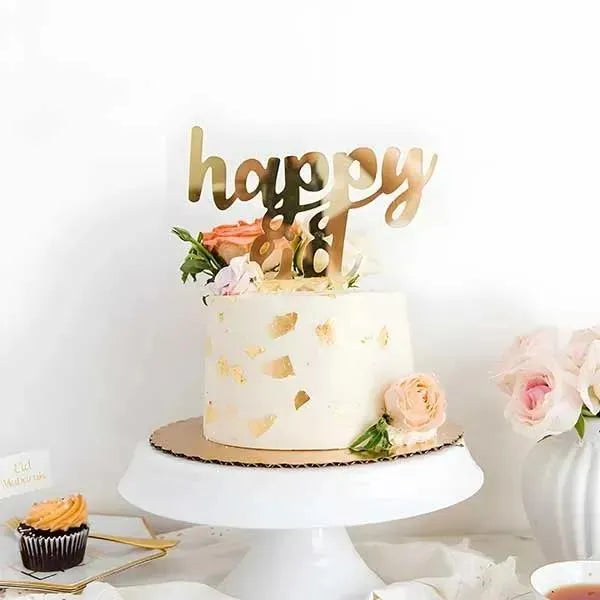 A decorative cake on a white pedestal, featuring a smooth, pale-colored frosting with gold leaf accents. Atop the cake is a golden acrylic topper that reads "happy eid" in a playful script. Surrounding the cake are fresh flowers in soft pink and green hues. In the foreground, there are additional desserts, including a cupcake with orange frosting, and a small card that says "Eid Mubarak." The background is simple and light, enhancing the festive presentation.