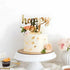 A decorative cake on a white pedestal, featuring a smooth, pale-colored frosting with gold leaf accents. Atop the cake is a golden acrylic topper that reads "happy eid" in a playful script. Surrounding the cake are fresh flowers in soft pink and green hues. In the foreground, there are additional desserts, including a cupcake with orange frosting, and a small card that says "Eid Mubarak." The background is simple and light, enhancing the festive presentation.