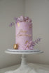 A beautifully decorated round cake sitting on a white cake stand. The cake is a soft lavender color and features a golden inscription that reads "Happy Anniversary." Adorning the top of the cake are edible decorations, including geometric candy pieces, purple and lavender flowers, and gold accents. A gold macaron is also placed at the base of the cake, completing the elegant design. The background is neutral, emphasizing the cake&