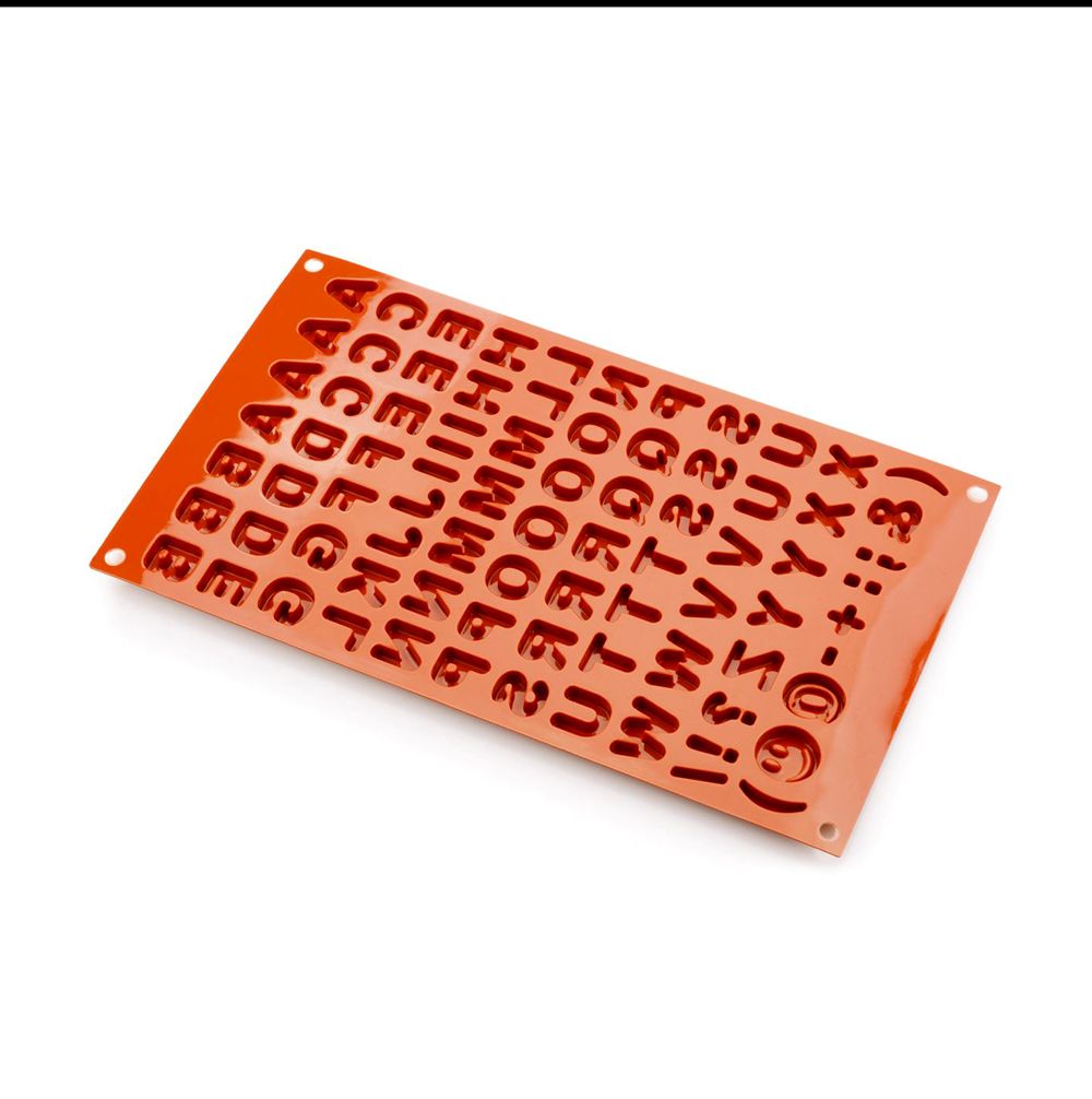 Red silicone mold with alphabet and numbers in reverse relief. The rectangular mold has rows of letters and symbols, conveying a playful tone.