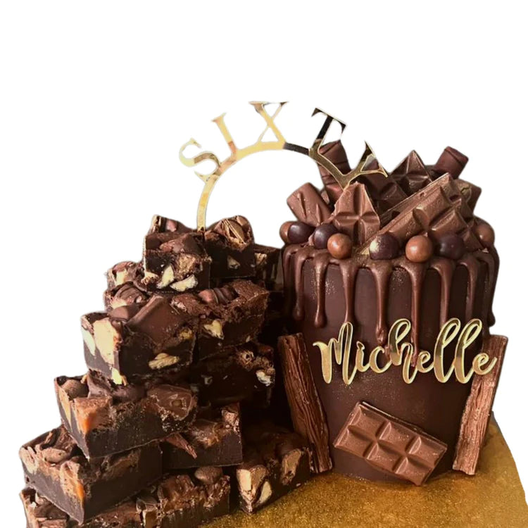 A chocolate cake with a glossy drip effect, topped with various chocolate pieces, round candies, and a decorative acrylic cake topper that reads &quot;SIXTY.&quot; The cake is positioned next to a stack of chocolate fudge pieces. The background is a golden surface, enhancing the festive appearance of the dessert.