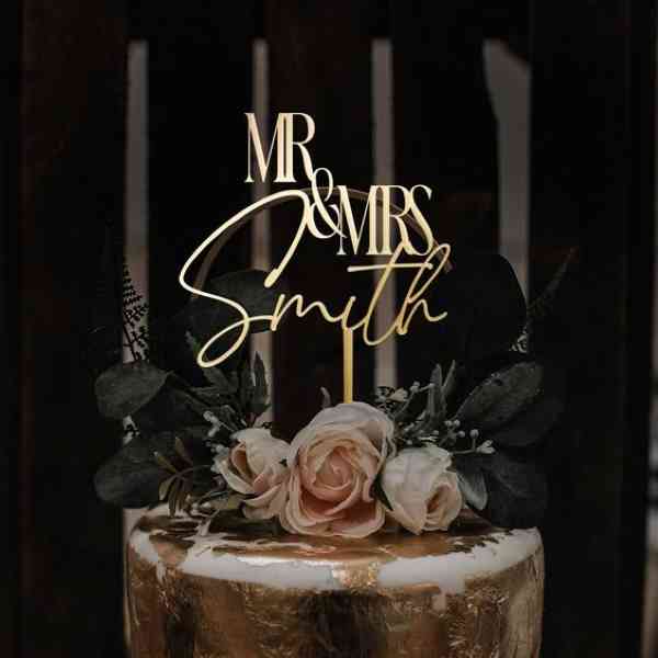 A cake topper featuring the phrase &quot;MR &amp; MRS&quot; in an elegant font, along with the name &quot;Smith.&quot; The topper is placed atop a tiered cake adorned with soft pink roses and greenery, including dark leaves. The cake has a gold finish, adding a touch of sophistication to the overall presentation. The background is dark, enhancing the visual impact of the cake and topper.