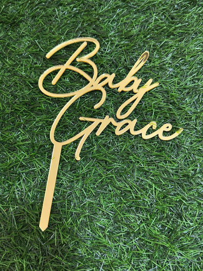 The image features a customized acrylic cake topper designed for celebrations. The topper is shaped in an elegant script font that reads "Baby Grace." It has a shiny gold finish and includes a tapered spike at the bottom for easy insertion into a cake. The background consists of green grass or faux grass, enhancing the visual presentation of the cake topper.