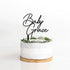 A round cake with a smooth, light-colored frosting is placed on a wooden cake stand. Atop the cake sits a customized acrylic cake topper featuring the words "Baby Grace" in an elegant, cursive font. The base of the topper is adorned with a decorative ring of green foliage. The background is plain and white, emphasizing the cake and topper.