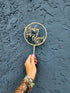 A hand is holding an acrylic cake topper that features the words "Baby in Bloom" inside a circular design. The topper includes decorative elements, such as flowers and leaves. The hand is adorned with multiple colorful bracelets, and the background is a textured dark blue wall.