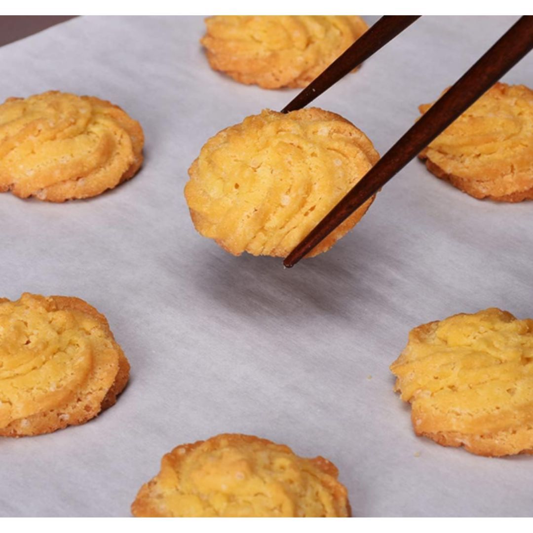 Golden brown cookies with a swirled design sit on parchment paper. A pair of chopsticks carefully lifts one cookie, highlighting its crispy texture.