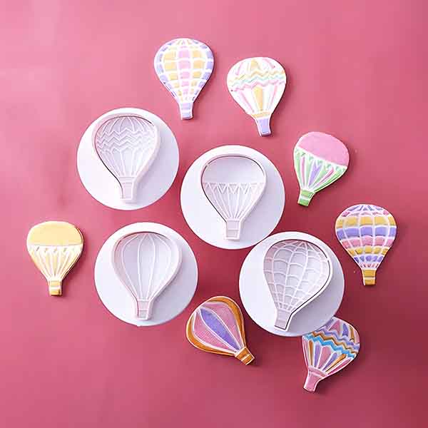 A collection of colorful hot air balloon-shaped cookies displayed on a pink background. The cookies feature various pastel designs and are surrounded by white circular plates. The arrangement includes four distinct hot air balloon shapes, with some cookies positioned upright and others laid flat, showcasing their decorative patterns.