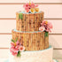 A two-tiered cake decorated to resemble bamboo, featuring realistic bamboo detailing along the sides. The cake is adorned with various colorful flowers, including pink and yellow blooms, and green leaf accents. The top tier has a light blue border, while the bottom tier has a textured white icing finish. The background features a simple wall, enhancing the cake&