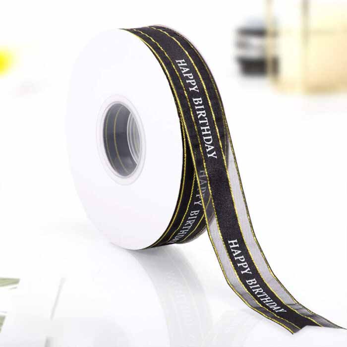 A roll of ribbon with a 2.5 cm width is displayed, featuring the text &quot;HAPPY BIRTHDAY&quot; printed in white on a black background. The ribbon has a shiny finish and is slightly curved, resting on a white spool. The background is blurred, suggesting an indoor setting or table surface.