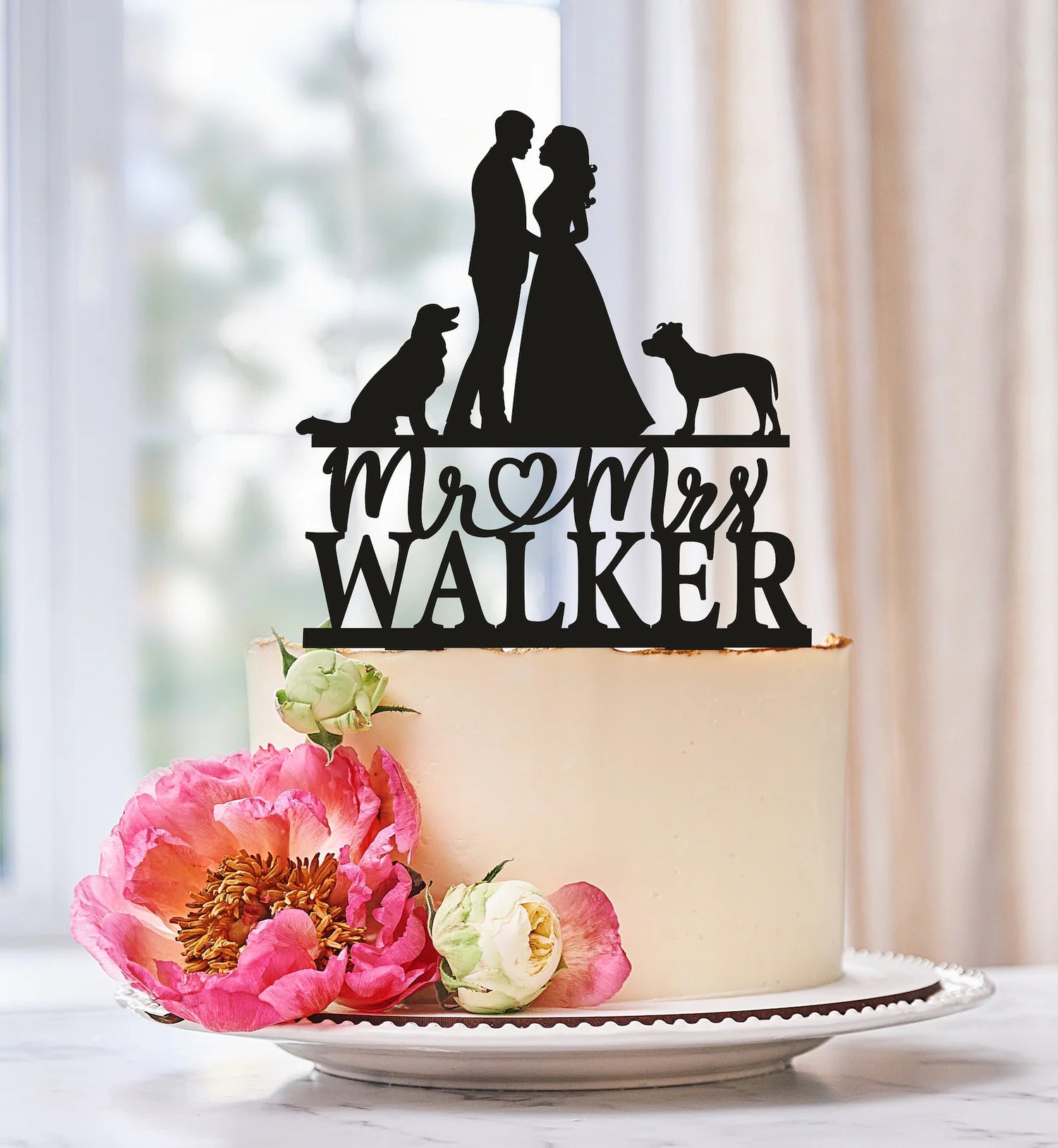A custom black acrylic cake topper featuring a silhouette of a couple standing together, with two dogs next to them. The text "Mr. &amp; Mrs. Walker" is prominently displayed below the couple. The topper is placed on a white cake adorned with pink peonies and smaller flowers, resting on a decorative plate against a softly blurred background.