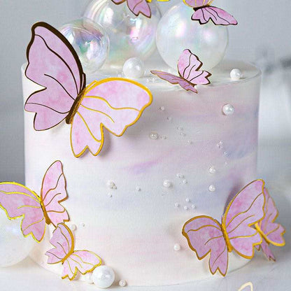 A decorative cake features a smooth, white surface with a subtle iridescent sheen. Multiple pink butterfly toppers, outlined in gold, are artfully arranged on the cake, some positioned on the sides and others on the top. The butterflies vary in size and orientation. Additionally, the cake is adorned with shiny, clear bubbles and small, round, white decorations that resemble pearls, enhancing its whimsical look.
