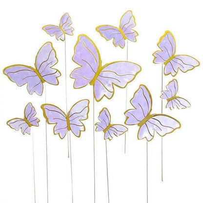 A collection of butterfly cake toppers in various sizes, featuring a soft lavender color with gold outlines. The butterflies are attached to thin white sticks, giving the appearance of them fluttering above a cake. There are a total of five butterflies in different orientations, some larger and some smaller, creating a whimsical and decorative effect.