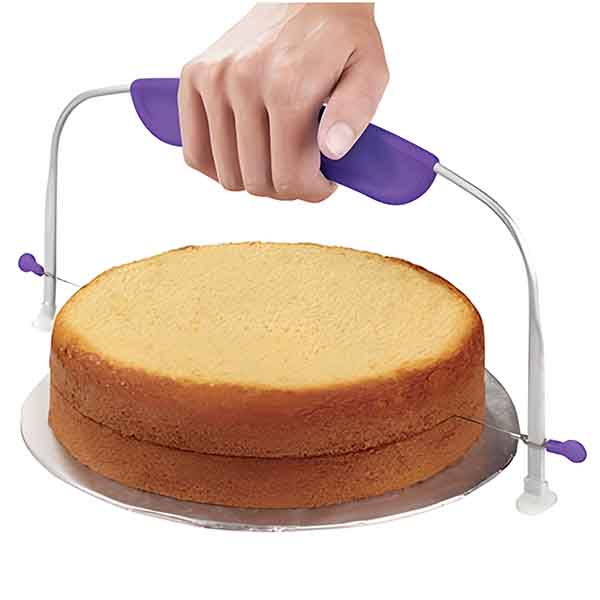 A cake leveler is shown in the image, featuring a person?s hand gripping the handle. The tool has a curved metal frame with two adjustable wires on either side. It is positioned above a round, beige cake placed on a silver cake board. The cake has a flat top and slightly rounded edges, indicating it is ready to be leveled. The handle is purple in color, providing a comfortable grip for use.