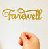 A person holds a gold glittery cake topper that spells "Farewell" in an elegant script. The topper is attached to a clear stick and is set against a plain white background.