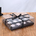 The image shows a clear cupcake box designed to hold 12 portions. The box features a transparent top with a decorative black ribbon tied around it, which has the inscription "Best Wishes for You." Inside, the base of the box is black and holds 12 circular cupcake holders. The box is resting on a wooden surface with a softly blurred background.